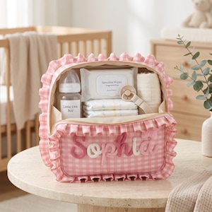May include: A pink and white gingham patterned storage bag with ruffled edges and the name Sophia embroidered in pink on the front. The open bag contains an 8oz baby bottle, a pack of sensitive wipes, two nappies, and a dummy with a clip.