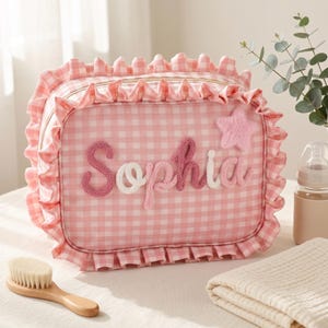 May include: A rectangular pink and white gingham toiletry bag featuring a ruffled border and a gold zipper. The name Sophia is written in raised, knitted script lettering across the front, accompanied by a small, fuzzy pink star appliqué.