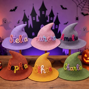 May include: Six pointed witch hats in purple, pink, grey, orange, yellow, and green. Each hat features a name in raised, colorful, knitted lettering: Stella, Aurora, Amelia, Sophia, Ethan, and Charlie. The hats have a textured, woven appearance.