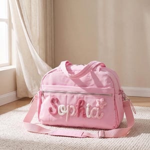 Könnte beinhalten: Eine hellrosa Reisetasche mit dem Namen Sophia in strukturierter, erhabener Schreibschrift auf der Vordertasche. Die Tasche verfügt über ein Hauptfach mit Reißverschluss, zwei Tragegriffe und einen verstellbaren Schultergurt mit silberfarbenen Beschlägen.