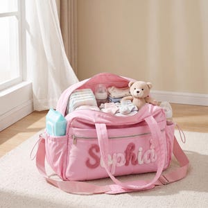 May include: A light pink diaper bag featuring the name Sophia in textured, raised lettering on the front pocket. The bag is open and filled with baby essentials, including folded clothing, a small teddy bear, and a clear plastic baby bottle.