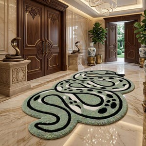 May include: A large, shaped area rug featuring a stylized snake design in shades of sage green, white, and black. The rug has a thick, tufted texture and follows the winding, coiled silhouette of a serpent, laid flat on a polished marble floor.