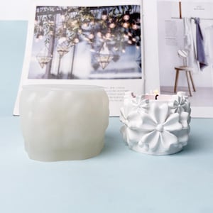 May include: A translucent white silicone mould with a textured surface and a finished white candle holder featuring a raised floral pattern. The candle holder has a cylindrical shape with multiple sculpted flower petals around the exterior.