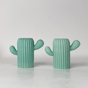 May include: A pair of identical light mint green cactus-shaped candle holders. Each holder features a cylindrical body with vertical ribbed textures and two small, curved arms extending from the sides, creating a symmetrical desert-inspired design.