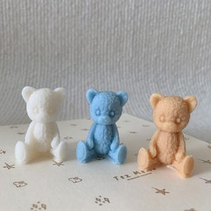 May include: Three small, seated teddy bear figurines in white, light blue, and peach colors. Each bear features a textured, fur-like surface and rounded ears. They are positioned on a cream-colored surface decorated with small brown star and rabbit icons.