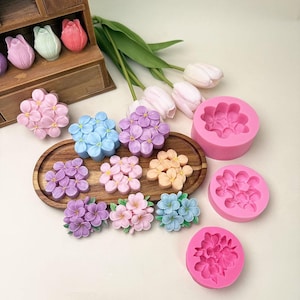 May include: A collection of silicone molds and finished flower-shaped resin or wax castings in various pastel colors including pink, blue, purple, and peach. The set includes three round pink molds and several floral pieces arranged on a wooden tray.