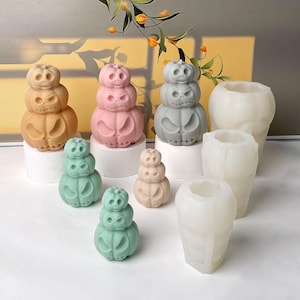 Può includere: Una collezione di figure impilate a forma di zucca in arancione, rosa, grigio e verde, insieme a stampi in silicone traslucidi. Ogni figura presenta tre livelli di volti intagliati con espressioni varie su piedistalli cilindrici bianchi.