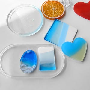 May include: A collection of resin coasters and trays in various shapes and colours, including a clear circular coaster, an orange slice coaster, a blue and white square coaster, a blue heart coaster, and an oval tray holding two small resin pieces.