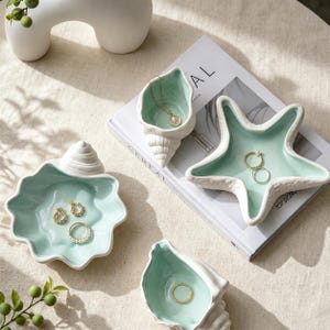 Könnte beinhalten: Ein Set aus vier keramischen Schmuckschalen in Form von Muscheln und einem Seestern, mit weißer Außenseite und hellminzgrünem Inneren. Die Schalen enthalten verschiedene goldfarbene Schmuckstücke, darunter Ringe, Ohrringe und eine zarte Halskette.