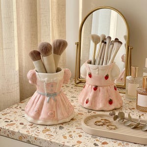 May include: Two ceramic holders shaped like dresses, one pink with a blue bow and floral details, the other pink with strawberry patterns. Both contain makeup brushes. A gold-framed mirror and a beige tray with gold rings are also present.