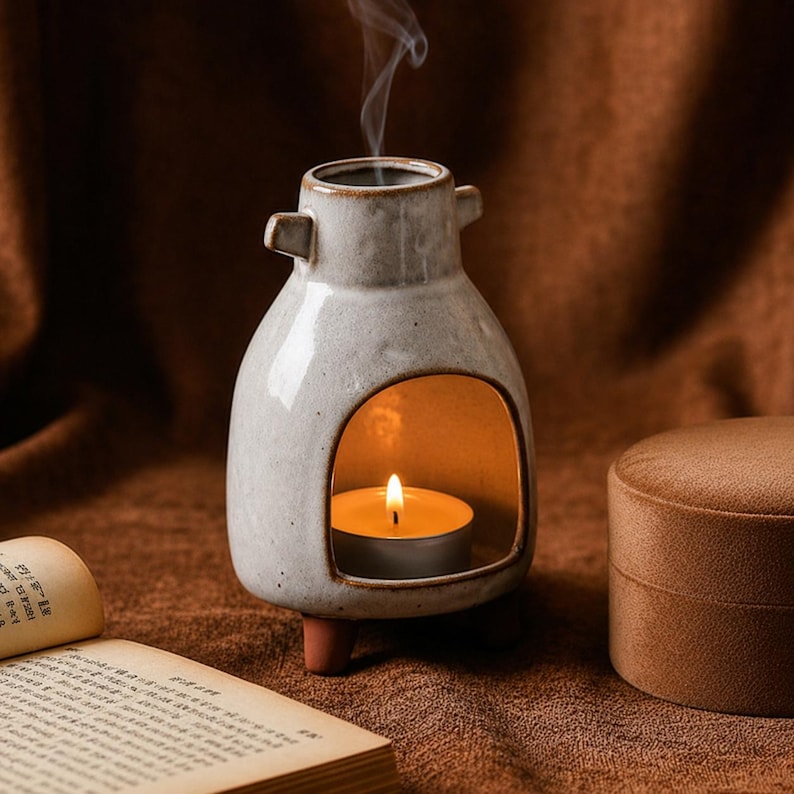 Może przedstawiać: Kremowy ceramiczny kominek do olejk&oacute;w z nakrapianym szkliwem, posiadający szeroki łukowaty otw&oacute;r u podstawy na świeczkę typu tealight. Naczynie ma dwa małe uchwyty po bokach, trzy kr&oacute;tkie brązowe n&oacute;żki oraz wąski otw&oacute;r u g&oacute;ry, z kt&oacute;rego unosi się dym.