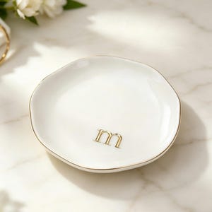 Peut inclure: Un petit vide-poche en céramique blanche de forme ronde avec un bord irrégulier et organique. Le plat présente un bord doré métallique et une lettre m minuscule dans une police serif dorée centrée près du bas de la surface intérieure.
