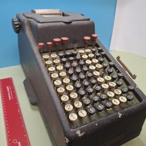 May include: An old black mechanical calculator featuring numerous numbered keys and red reset buttons. The calculator has a rectangular shape with a slightly angled top and a paper output slot. A red ruler is on the left.