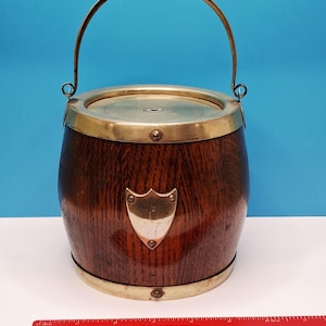 May include: A cylindrical wooden barrel with a brass rim, base, and arched handle. The barrel features a brass shield-shaped plaque on the front and a circular brass lid. A red ruler is positioned in front, showing a width of approximately 15 cm.