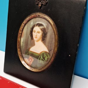 May include: A square black wall plaque featuring an oval portrait of a person with dark hair wearing a green dress and holding a small book. The plaque has a metallic acorn-shaped hanger at the top and is shown next to a red ruler for scale.