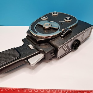 May include: A black Quarz 8mm movie camera with a textured finish, a side-mounted grip, and a silver trigger. The camera features a circular top dial, a lens, and is positioned next to a red ruler showing a length of approximately 20 cm.