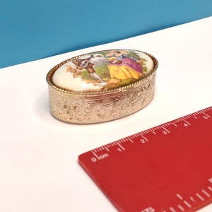 May include: An oval metal trinket box with an embossed floral pattern on the sides and a lid featuring a colourful painted scene of two figures. A red plastic ruler is placed next to the box, showing a length of approximately 6.5 cm.