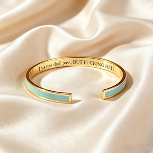 Puede incluir: Un brazalete dorado con una incrustación de esmalte azul claro en el exterior. La superficie interior está grabada con el texto en negro, This too shall pass, BUT FUCKING HELL. El brazalete tiene un diseño abierto con un acabado pulido.
