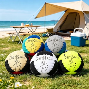 Puede incluir: Una colección de seis pelotas Dirtbag coloridas, cada una con un esquema de color único y el logotipo de Dirtbag. Las pelotas están dispuestas sobre una superficie de césped cerca de una playa, con una tienda de campaña y una mesa de picnic al fondo.
