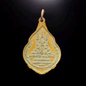 May include: A teardrop-shaped gold-toned metal pendant featuring intricate embossed Thai script and sacred geometric symbols on its surface. The pendant has a small circular bail at the top for attachment and a textured, weathered finish.