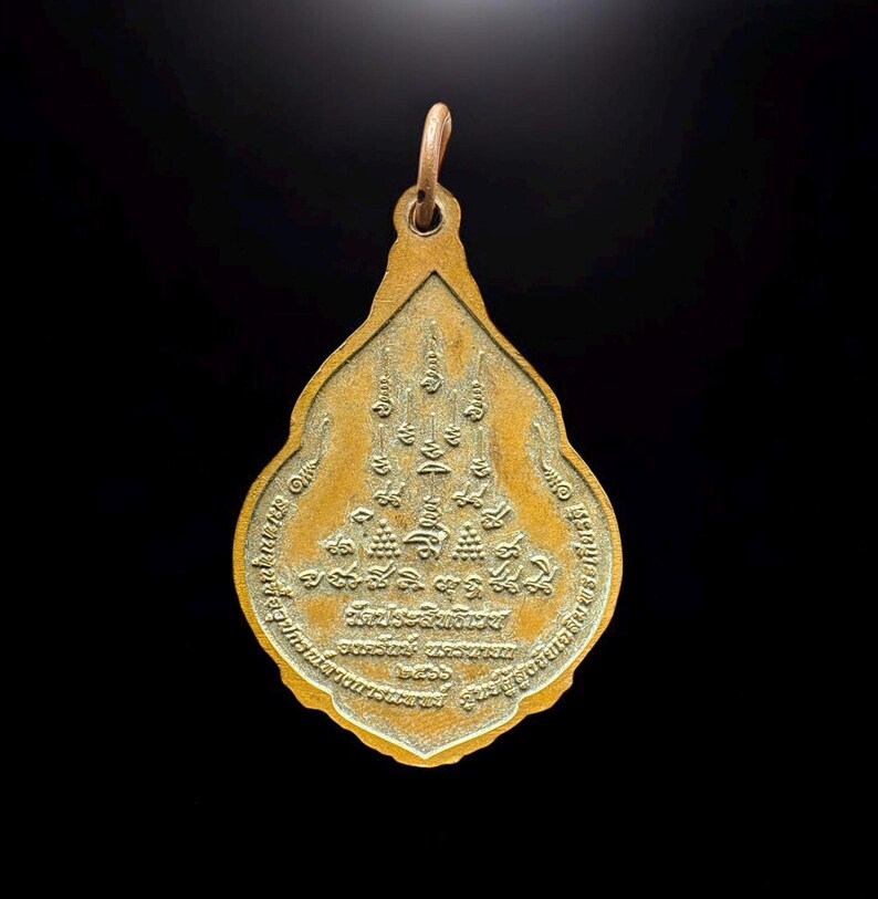 May include: A teardrop-shaped gold-toned metal pendant featuring intricate embossed Thai script and sacred geometric symbols on its surface. The pendant has a small circular bail at the top for attachment and a textured, weathered finish.
