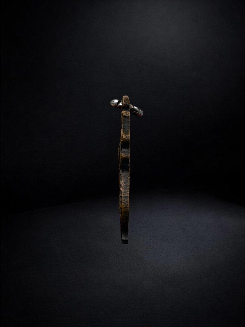 May include: A side profile view of a dark brown, textured, vertical pendant with a weathered finish. The object features a metallic silver-toned jump ring attached to the top, providing a clear view of its slim, elongated shape and rugged surface.