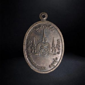 Puede incluir: Un colgante de metal de tono bronce con forma ovalada y una peque&ntilde;a anilla circular en la parte superior. La superficie presenta inscripciones en tailand&eacute;s y marcas simb&oacute;licas en relieve dispuestas en un patr&oacute;n circular alrededor de un emblema central.