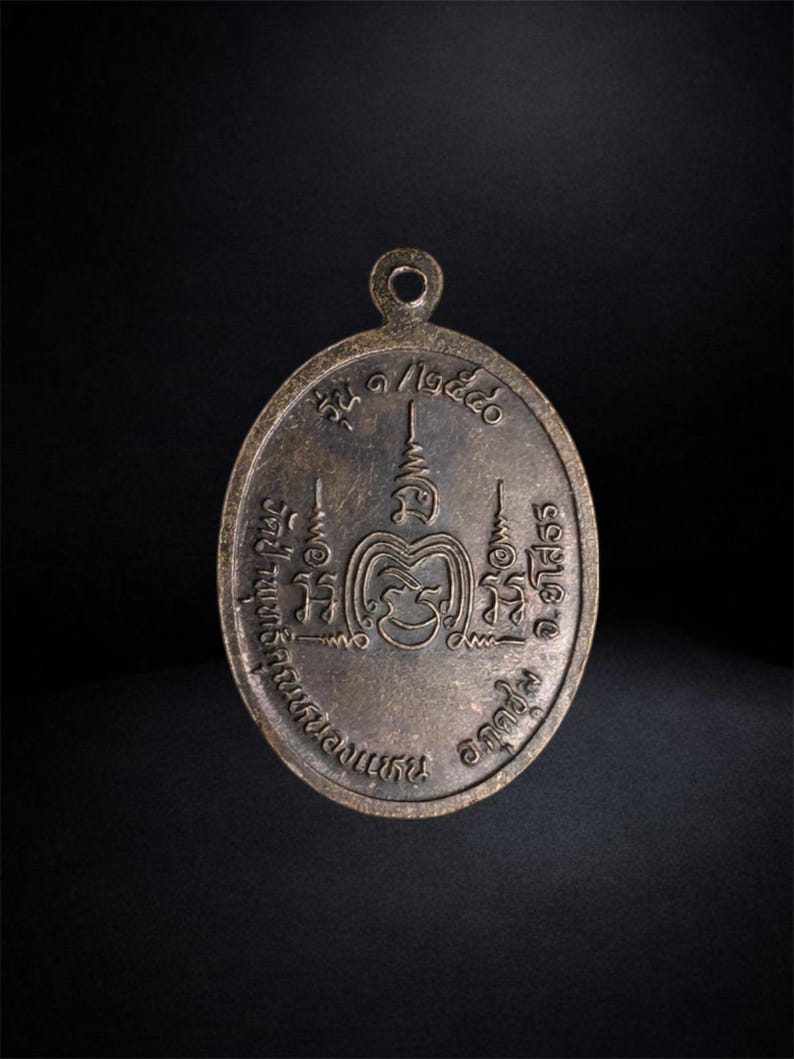 Puede incluir: Un colgante de metal de tono bronce con forma ovalada y una peque&ntilde;a anilla circular en la parte superior. La superficie presenta inscripciones en tailand&eacute;s y marcas simb&oacute;licas en relieve dispuestas en un patr&oacute;n circular alrededor de un emblema central.