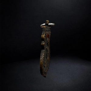 May include: A dark, weathered metallic pendant with a textured, uneven surface and a prominent raised relief design. The object features a silver-toned metal loop at the top for suspension, showcasing a deep bronze and black patina throughout its body.