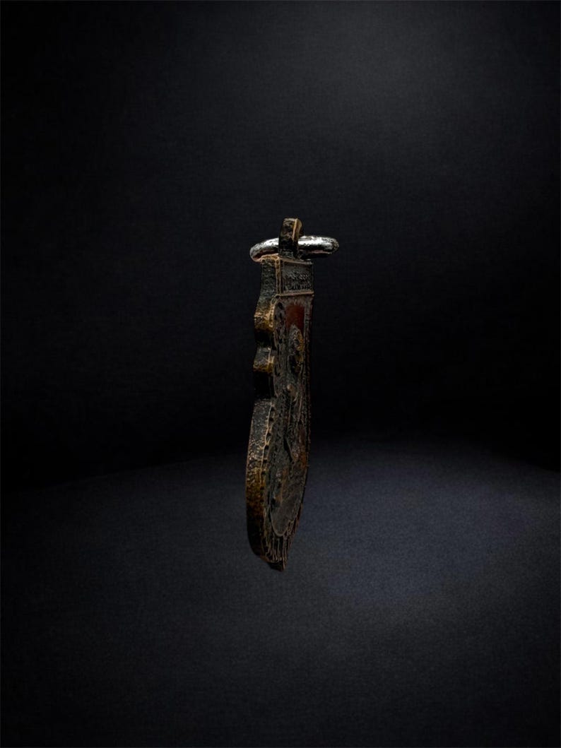 May include: A dark, weathered metallic pendant with a textured, uneven surface and a prominent raised relief design. The object features a silver-toned metal loop at the top for suspension, showcasing a deep bronze and black patina throughout its body.