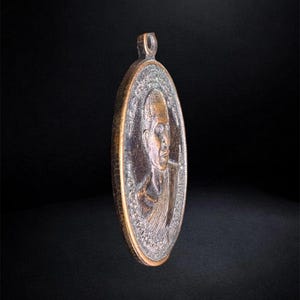 Puede incluir: Un colgante ovalado de metal en tono bronce que presenta un retrato en relieve de una persona con la cabeza rapada. El colgante tiene un borde texturizado y una peque&ntilde;a anilla en la parte superior para colgarlo. La superficie muestra un acabado met&aacute;lico envejecido.
