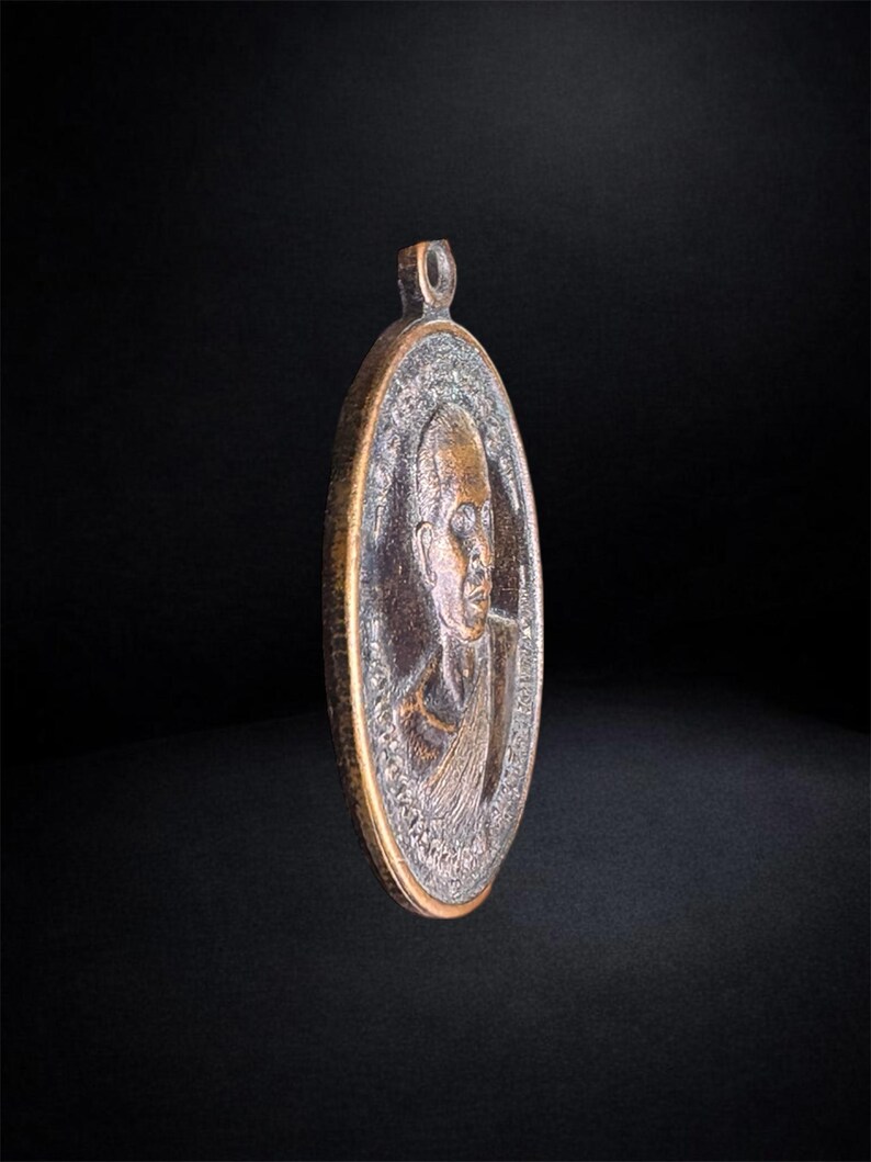 Puede incluir: Un colgante ovalado de metal en tono bronce que presenta un retrato en relieve de una persona con la cabeza rapada. El colgante tiene un borde texturizado y una peque&ntilde;a anilla en la parte superior para colgarlo. La superficie muestra un acabado met&aacute;lico envejecido.