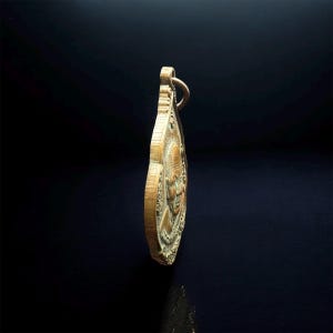 May include: A bronze-toned metal pendant shaped like a teardrop, featuring a relief portrait of a person on its surface. The pendant has a small circular loop at the top for attachment and displays a textured, aged finish with visible metallic details.