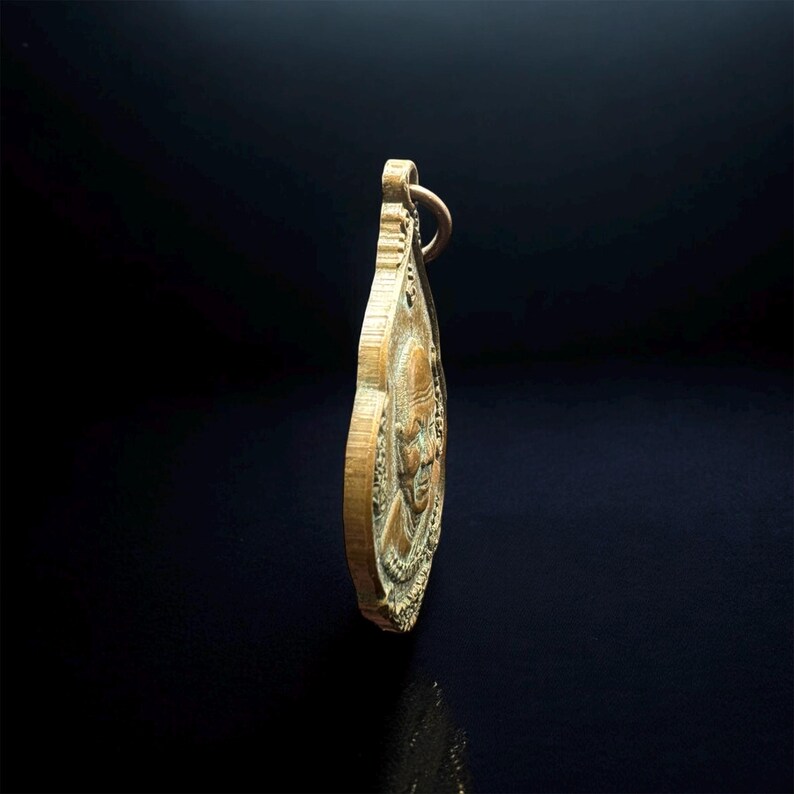 May include: A bronze-toned metal pendant shaped like a teardrop, featuring a relief portrait of a person on its surface. The pendant has a small circular loop at the top for attachment and displays a textured, aged finish with visible metallic details.
