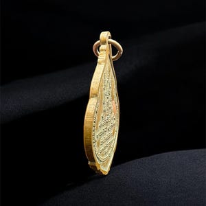May include: A gold-toned metal pendant shaped like a teardrop or leaf, featuring intricate embossed detailing on its surface. The pendant has a small circular loop at the top for attachment and displays a textured, relief-style design throughout.