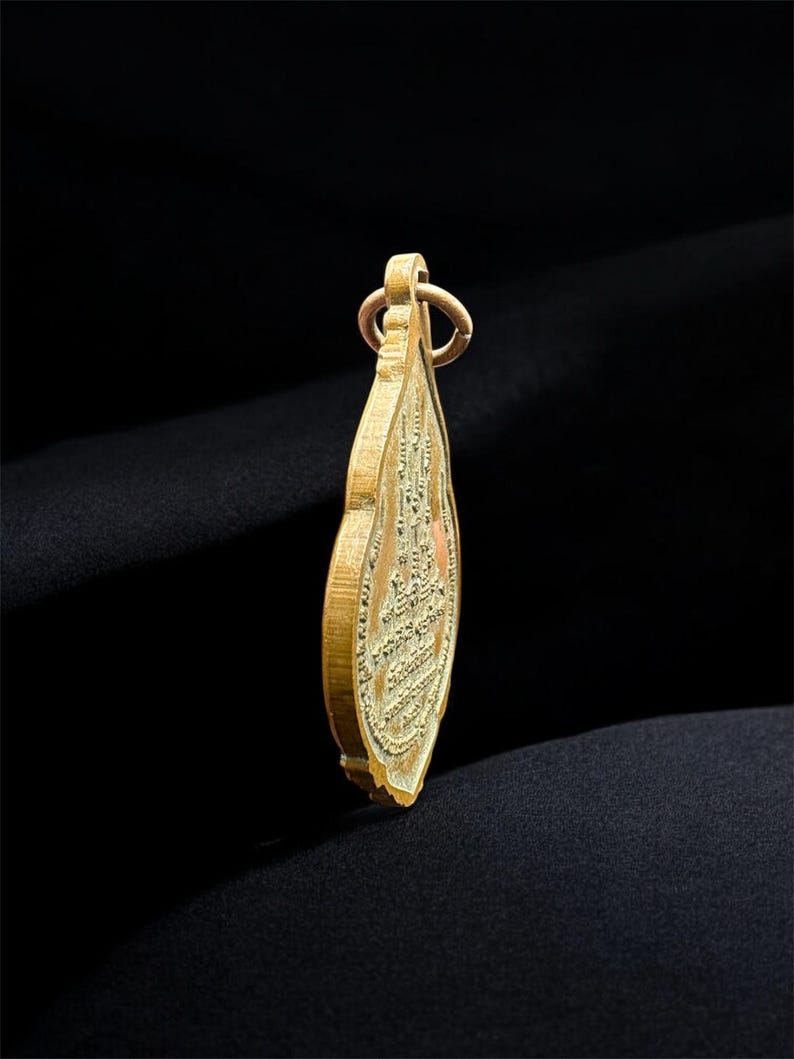 May include: A gold-toned metal pendant shaped like a teardrop or leaf, featuring intricate embossed detailing on its surface. The pendant has a small circular loop at the top for attachment and displays a textured, relief-style design throughout.