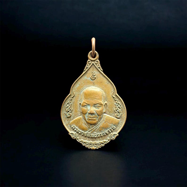 May include: A bronze-toned teardrop-shaped pendant featuring a relief portrait of an elderly person with a shaved head. The pendant has an ornate border with decorative scrollwork and Thai script embossed along the bottom edge and a bail at the top.