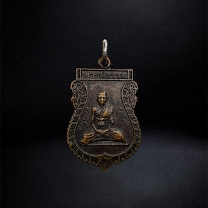 May include: A bronze-toned metal pendant featuring a relief of a seated figure in robes. The shield-shaped charm has ornate decorative borders, Thai script text at the top and sides, and a silver-colored metal jump ring attached to the top loop.
