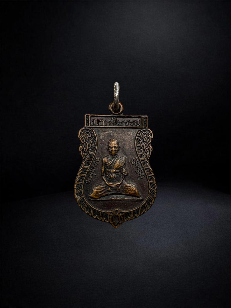 May include: A bronze-toned metal pendant featuring a relief of a seated figure in robes. The shield-shaped charm has ornate decorative borders, Thai script text at the top and sides, and a silver-coloured metal jump ring attached to the top loop.
