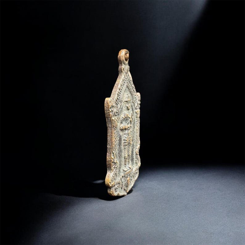 May include: A bronze-toned metal pendant featuring a detailed relief of a standing figure in traditional attire. The pendant has an elongated, pointed shape with intricate decorative borders and a small circular loop at the top for suspension.