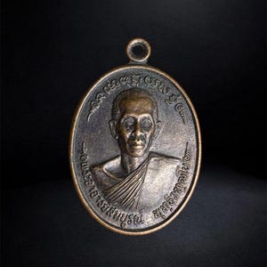 Puede incluir: Un colgante de metal de tono bronce con forma ovalada que presenta un retrato en relieve de una persona con t&uacute;nicas tradicionales. La superficie est&aacute; grabada con caracteres en tailand&eacute;s que rodean el retrato y tiene una peque&ntilde;a anilla circular.