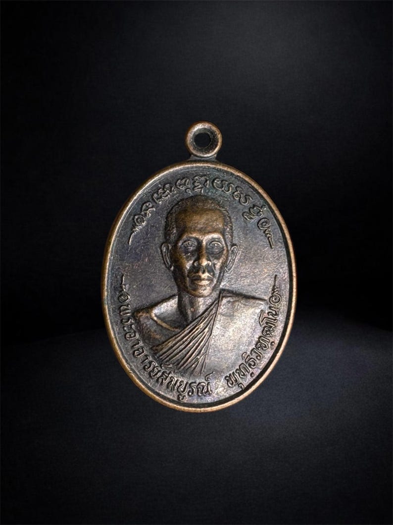 Puede incluir: Un colgante de metal de tono bronce con forma ovalada que presenta un retrato en relieve de una persona con t&uacute;nicas tradicionales. La superficie est&aacute; grabada con caracteres en tailand&eacute;s que rodean el retrato y tiene una peque&ntilde;a anilla circular.