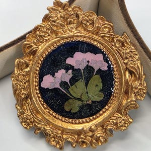 May include: A round brooch featuring a gold-colored ornate frame with intricate floral relief details. The center contains a dark, glittery blue resin inlay embedded with pressed pink flowers and green leaves, creating a botanical display.