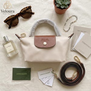 May include: Cream-colored handbag with a brown flap and handle, featuring a gold button closure. The bag is accompanied by a brown shoulder strap, sunglasses, perfume, and accessories. The bag has the brand name "Longchamp" on the flap.
