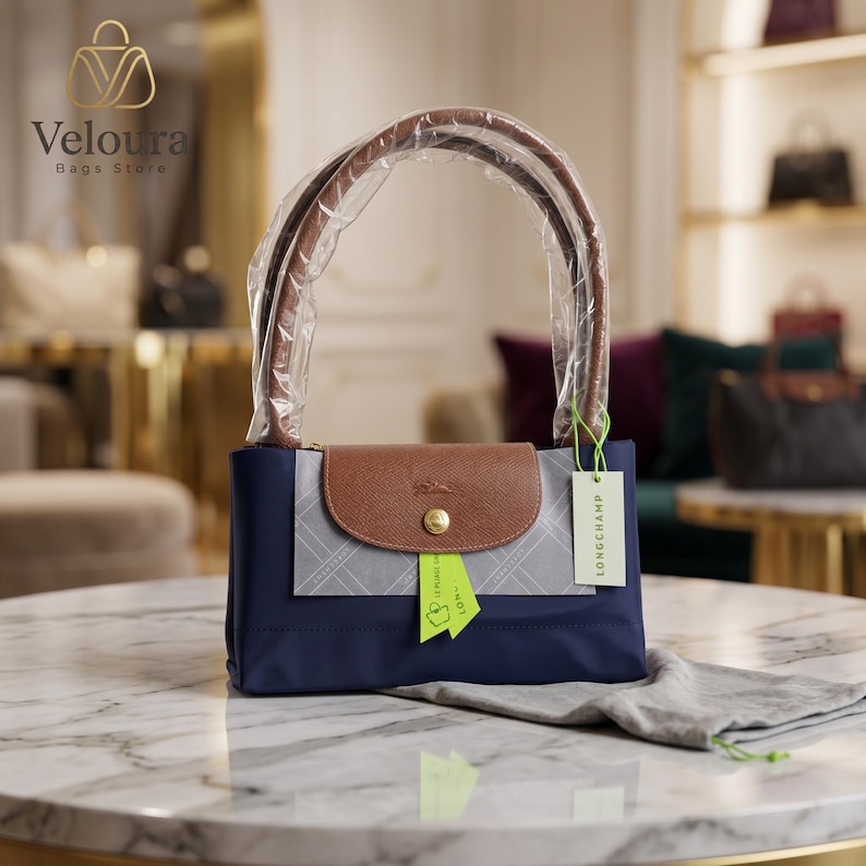 May include: A navy blue handbag with a brown leather flap and handles. The bag has a rectangular shape and a gold-colored button closure. A tag with the brand name "Longchamp" is attached.
