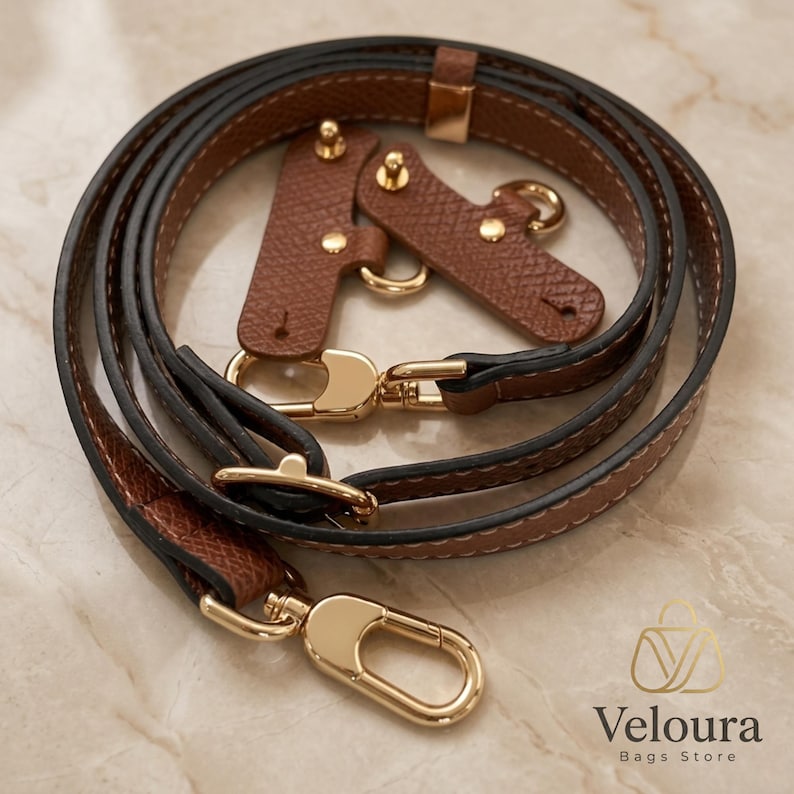 May include: A brown leather bag strap with gold-colored hardware. The strap is made of two layers of leather, with a darker brown on the outside and a lighter brown on the inside. The strap has gold-colored clasps and connectors.