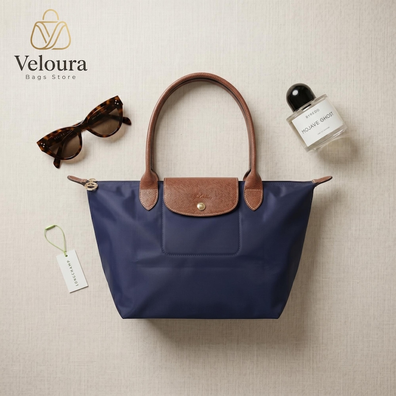 May include: Navy blue tote bag with brown leather handles and flap closure. Tortoiseshell sunglasses and a perfume bottle are next to the bag. The bag has a gold zipper and a small tag.