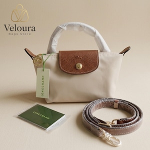 May include: A beige handbag with a brown leather flap and handle. The bag has a gold-colored clasp and a detachable brown strap. A tag that says "LONGCHAMP" hangs from the bag. A small green card with the brand name is also included.