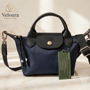 Puede incluir: Bolso de mano azul marino con detalles en cuero negro, un asa superior y una correa para el hombro desmontable. El bolso tiene un cierre de solapa con un botón dorado y una pequeña etiqueta. El bolso es de Veloura Bags Store.