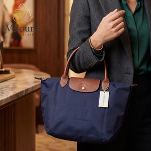 May include: A navy blue tote bag with brown leather handles and a flap closure. The bag has a rectangular shape and a small front pocket. A tag with the brand name is attached to the bag.